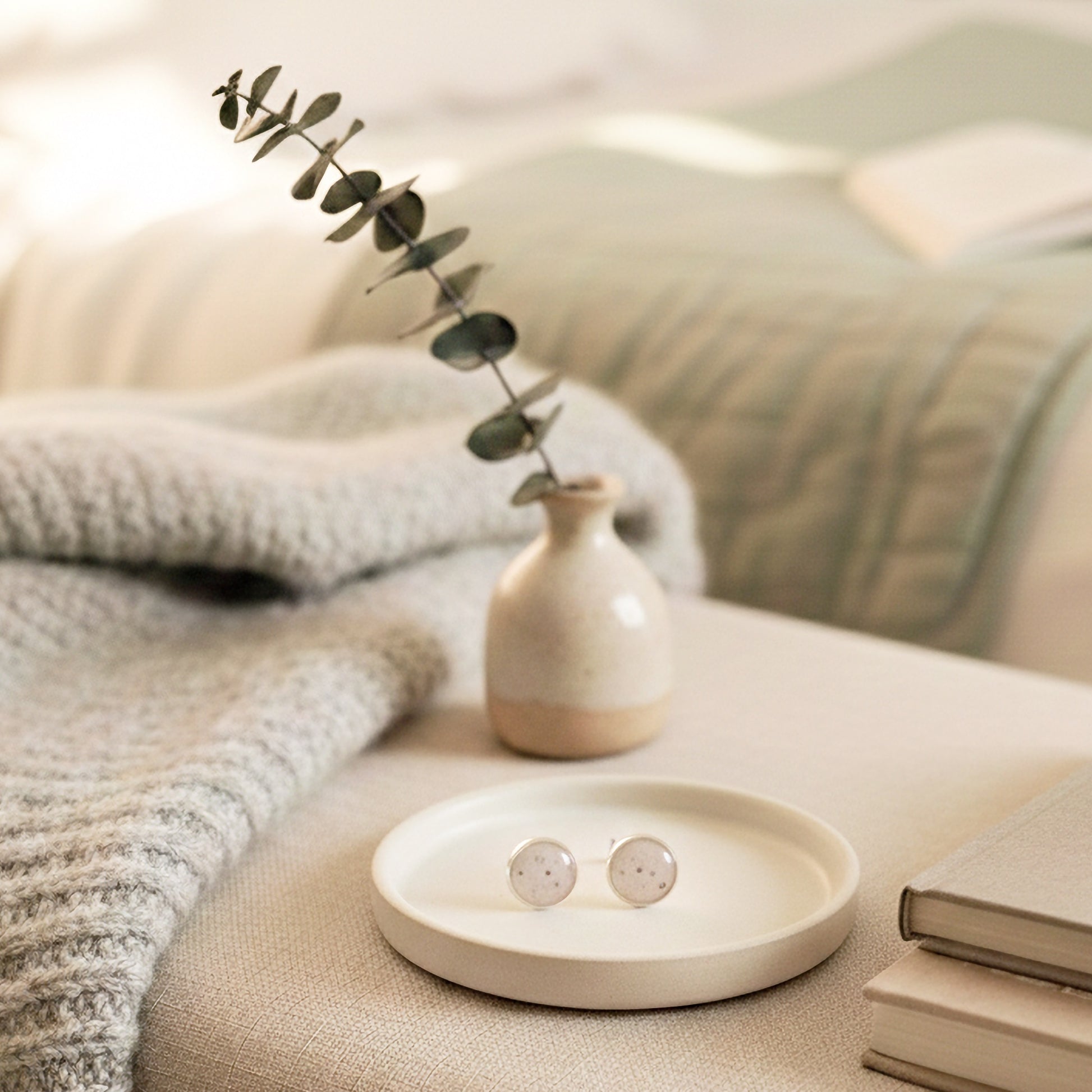 earrings in ceramic dish, vase with plant, and book on a soft surface