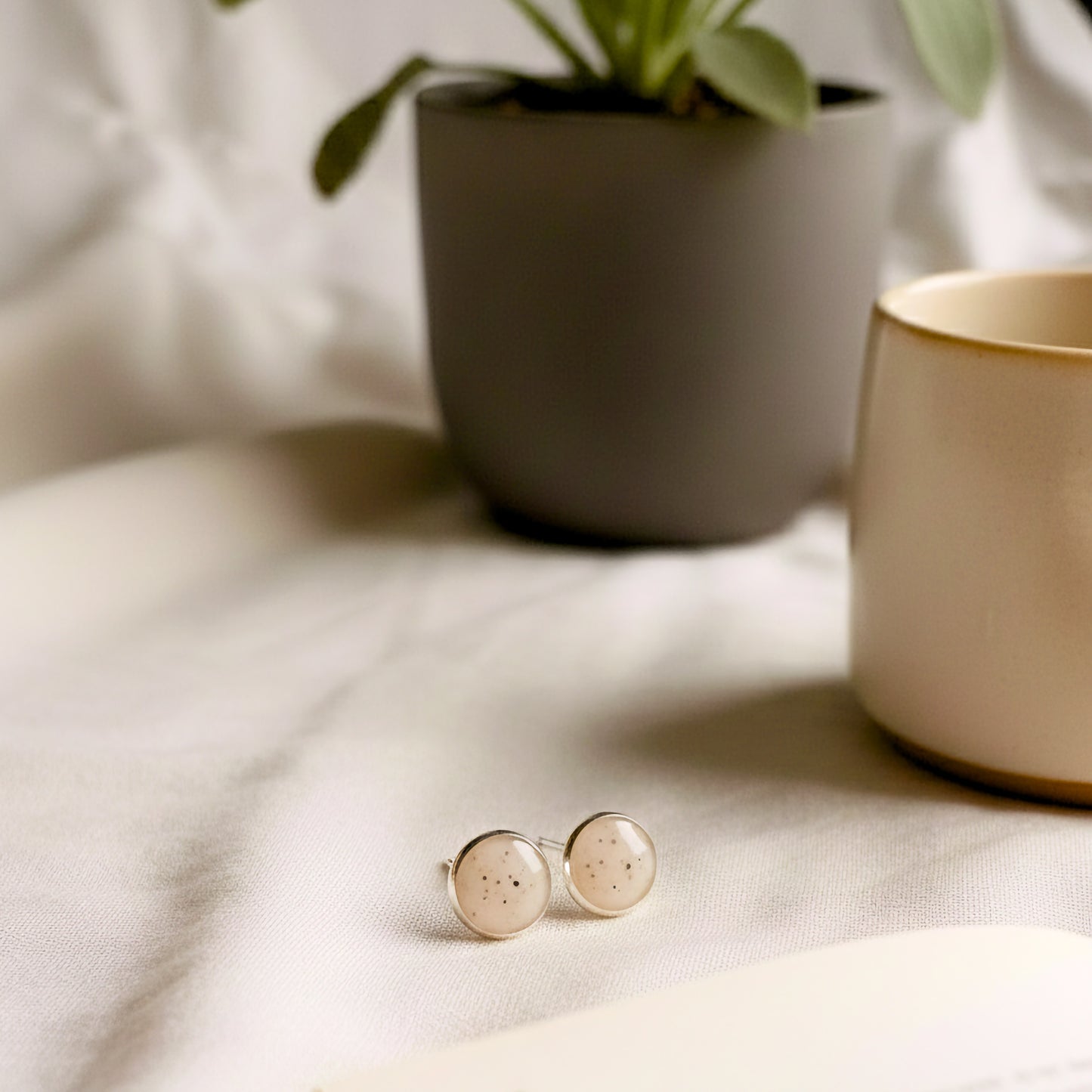 Pair of round earrings on a light fabric surface with a plant and mug in the background