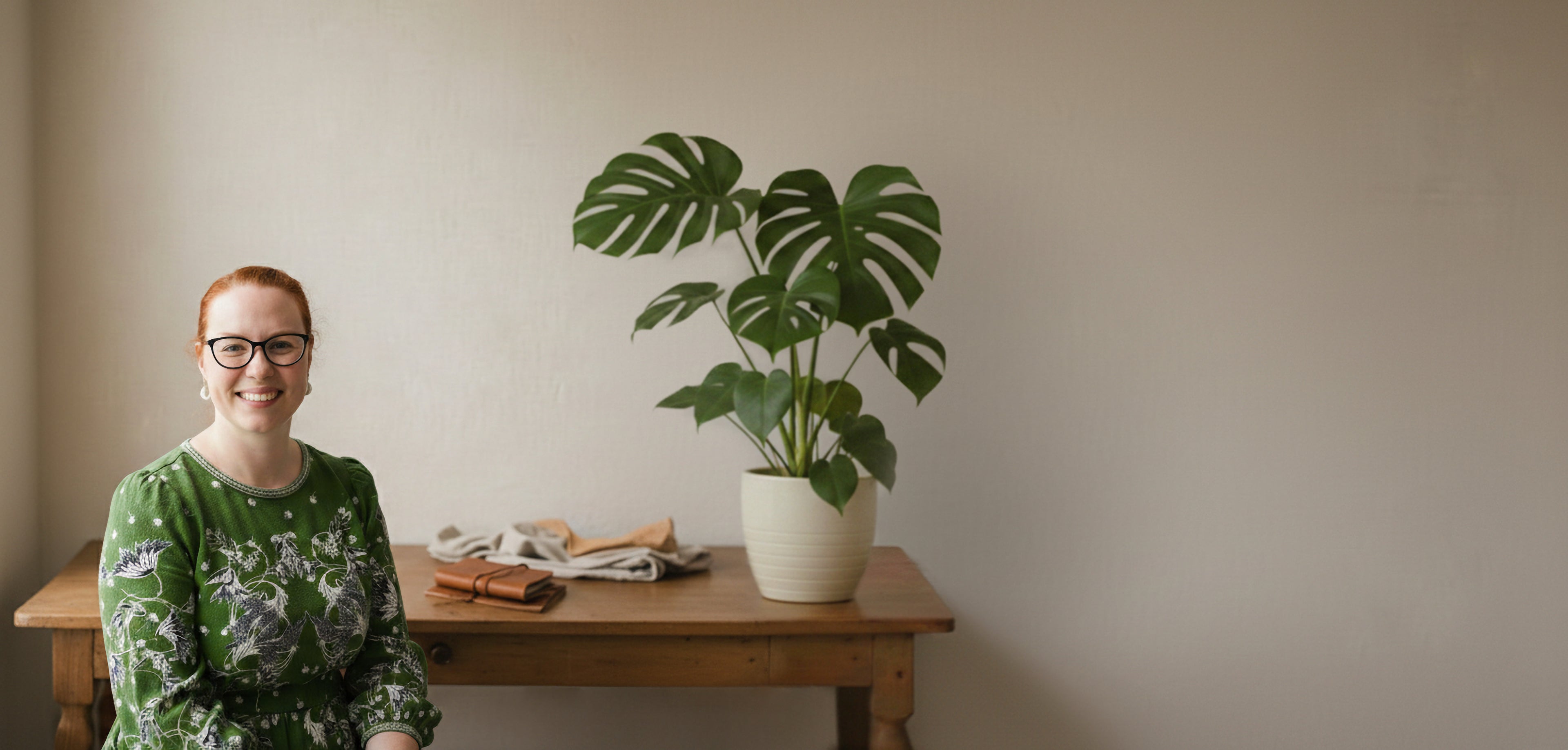 Woman sitting at a wooden table with a potted plant against a plain wall
