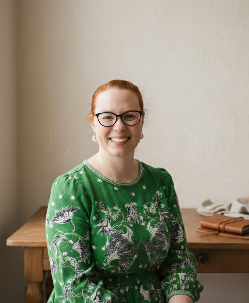 Woman wearing glasses and a green patterned dress sitting at a wooden table.