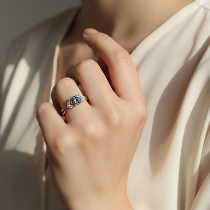 Hand wearing a ring with a blue-green moissanite gemstone against a neutral background